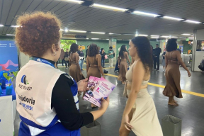 Mulher distribuindo panfletos na estação, e uma apresentação de ballet ao fundo